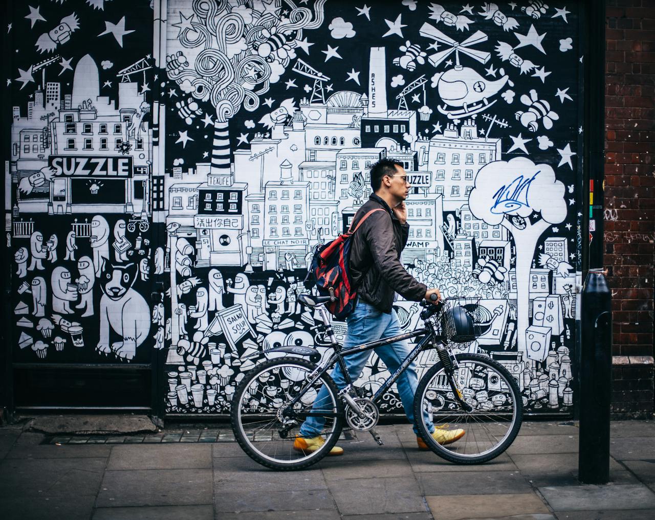 about-img Man walking a bicycle past a detailed urban graffiti mural in a city setting.